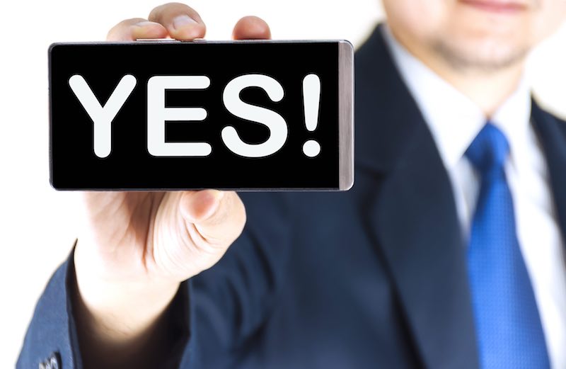 YES!, word on mobile phone screen in blurred young businessman hand over white background, business concept